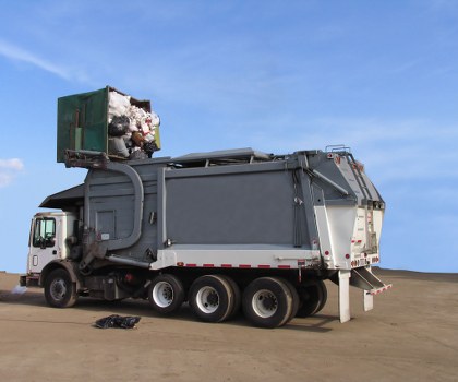 A large commercial waste collection truck with a white cab and a dark grey waste container, parked on an open paved area under a clear blue sky. The waste container is filled with various types of rubbish, including white bags and other debris, with the top opening revealing the contents and some waste visible at the edges. The truck is positioned horizontally across the image, showing multiple sets of black rubber tyres on both sides. Behind the truck, the landscape appears to be flat and open, with no other objects or structures visible nearby. The scene suggests a routine rubbish removal operation, possibly in a commercial or urban setting within Chelsea, as illustrated by the context of Commercial Waste Removal Chelsea. The overall environment is well-lit with natural daylight highlighting the textures of the waste and the metallic finish of the vehicle, aligning with professional rubbish collection services in the local area.