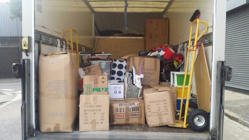 Inside a partially opened commercial waste removal van, there are various displaced cardboard boxes of different sizes, some sealed with packing tape and others open, revealing contents like paper and plastic waste. The boxes are stacked and arranged across the floor and along the back wall of the vehicle, which has a wooden interior paneling and a metallic ceiling with some visible hardware. Two yellow hand trucks with black wheels are positioned on either side of the cargo area, leaning against the sides, and one of them has a small red and black object on it. The floor of the van is cluttered with the cardboard containers, and the surrounding environment suggests a roadside or industrial area, with a grey shuttered door partially visible on the right. The scene indicates a waste collection or rubbish removal operation by Commercial Waste Removal Chelsea, with a focus on disposing of cardboard and general waste materials typical of commercial rubbish clearance in the Chelsea area, inferred from the context of the webpage.