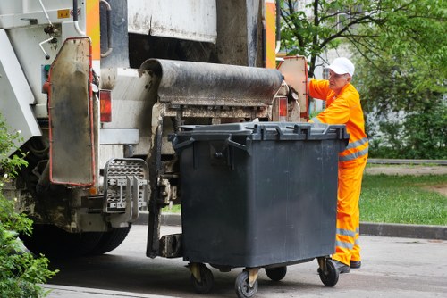 A waste collection vehicle with a large rotating drum at the rear is positioned outdoors on a paved area, adjacent to a grassy verge and some green trees. The vehicle is painted in neutral tones with visible metal and plastic components, and appears to be in operational condition. A worker dressed in high-visibility orange safety clothing, including a jacket, trousers, and a white safety helmet, is standing on the right side of the image, actively engaging with a black plastic rubbish bin placed near the vehicle. The bin has a smooth surface, a sturdy lid, and is mounted on small wheels. The worker is holding the bin’s lid or gripping its side, preparing to empty it into the vehicle’s hopper. The background suggests a suburban or urban environment, typical of commercial waste removal services in Chelsea, with overcast lighting and greenery providing contextual environmental detail. The scene captures a moment of rubbish collection consistent with professional waste management operations focused on local rubbish disposal.