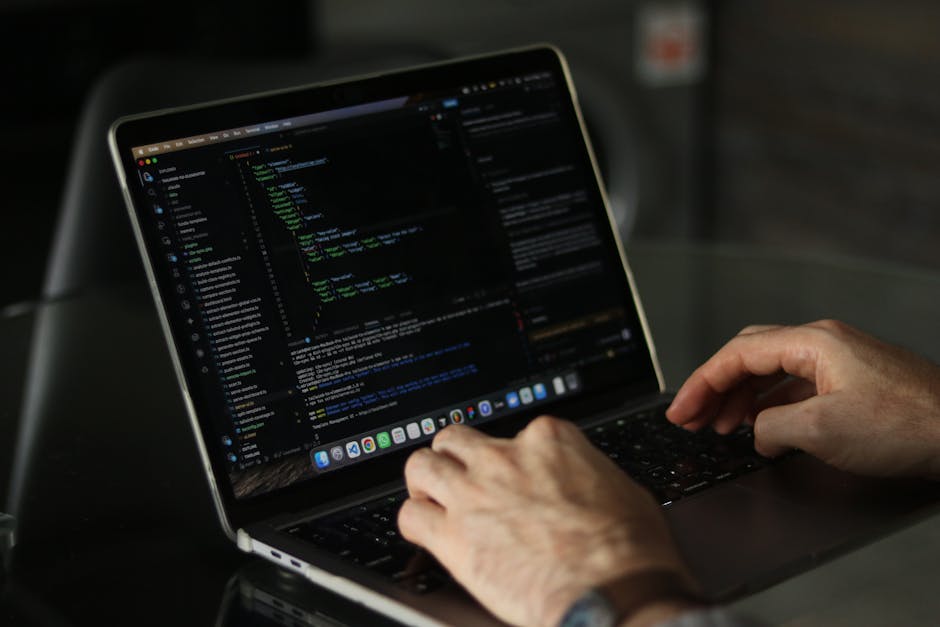A black laptop computer with a red backlit keyboard sits on a dark, smooth surface, with its screen displaying lines of colorful, blurred code or digital text, suggesting programming or hacking activi