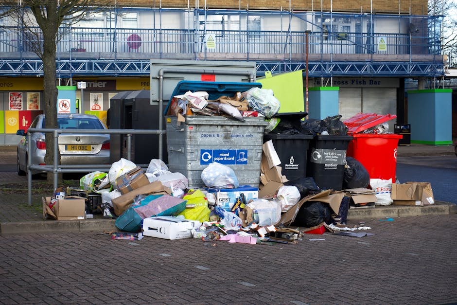 A large collection of overflowing rubbish bins and scattered waste bags located on a paved sidewalk in front of a commercial building. The waste includes black and clear plastic trash bags, flattened 