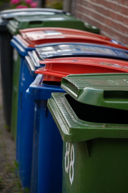 Five large black wheeled rubbish bins with yellow lids are positioned in a straight row on a concrete surface against a plain, pale grey wall. Each bin has a white oval sticker with black text, and th