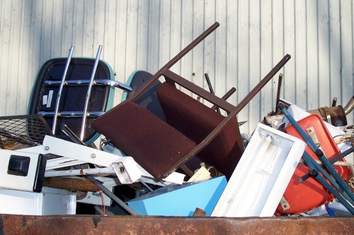 A large pile of assorted discarded items resting on a surface outdoors, including a dark blue plastic chair with metal legs, a brown wooden chair with a fabric seat in the foreground, a white plastic window frame, and a red plastic container with a handle. There are also metal and plastic components, some partially obscured, and the background consists of vertically aligned light-colored corrugated metal sheeting. The rubbish appears to be a mixture of furniture, fittings, and miscellaneous waste, indicative of an industrial or commercial disposal site. The image portrays a cluttered arrangement typical of waste removal operations in Chelsea, with various textures such as smooth plastics, painted surfaces, and rough metal. Ambient lighting suggests natural daylight, highlighting the details of the objects and their materials. As part of a professional rubbish removal service, Commercial Waste Removal Chelsea manages such waste collection, ensuring environmental standards are met while clearing commercial or domestic debris from the area near postal code SW3.