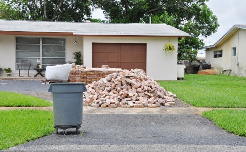 The image shows a pile of light pinkish-brown bricks and rubble, which appear to be construction or demolition waste, stacked on a paved driveway in front of a beige, single-story residential house with a brown garage door. To the left of the pile, a grey plastic waste bin with a dark lid is positioned on the driveway. The house features a large window with white frames and blinds, a small outdoor seating area with a black chair, and some bushes or plants near the wall. The surrounding area includes a well-maintained green lawn on both sides of the driveway with neatly cut grass, and the background features additional trees and a neighboring building partially visible to the right. The scene is outdoors during daylight, under overcast or partly cloudy skies. This setup exemplifies typical rubbish removal tasks handled by companies like Commercial Waste Removal Chelsea, serving areas such as Chelsea or nearby London postcodes, ensuring effective clearance of construction debris from residential properties.