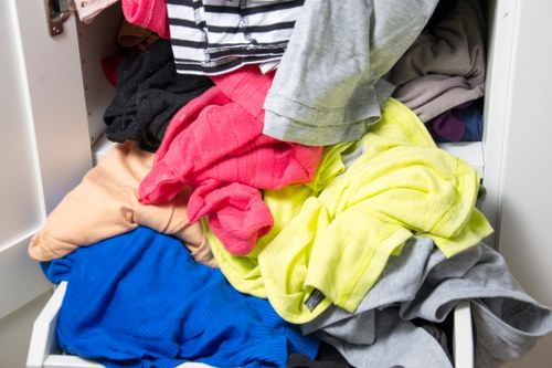 A close-up view of an open metal storage drawer or compartment filled with a disorganized assortment of various clothing items, including brightly coloured T-shirts, sweatshirts, and pants in shades of red, blue, yellow, grey, and beige. The clothing appears to be casual and mixed, with some garments folded while others are crumpled or stacked in a haphazard manner. The interior environment features a light-coloured metal finish, suggesting it may be part of a laundry or storage unit. The scene is well-lit, highlighting the different textures and colours of the fabrics, with no other objects or background elements visible that would suggest a specific location. The image does not depict any rubbish removal activity but shows clothing that could be destined for disposal, donation, or collection, fitting within the context of waste management and clearance services offered by Commercial Waste Removal Chelsea in the Chelsea area, near postcode SW3.