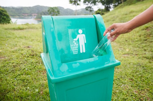 A person’s hand placing a clear plastic bottle into a bright turquoise wheelie bin with a white graphic of a person throwing waste into a bin, positioned outdoors on a grassy area near a body of water, with trees and cloudy sky in the background. The bin is situated on a paved surface, and the scene suggests a waste disposal or recycling activity typical of municipal or commercial rubbish collection in Chelsea, with natural greenery and a peaceful environment. The overall setting emphasizes the importance of proper waste separation and removal, relevant to services offered by Commercial Waste Removal Chelsea in the local borough, with the focus on environmentally conscious rubbish disposal.